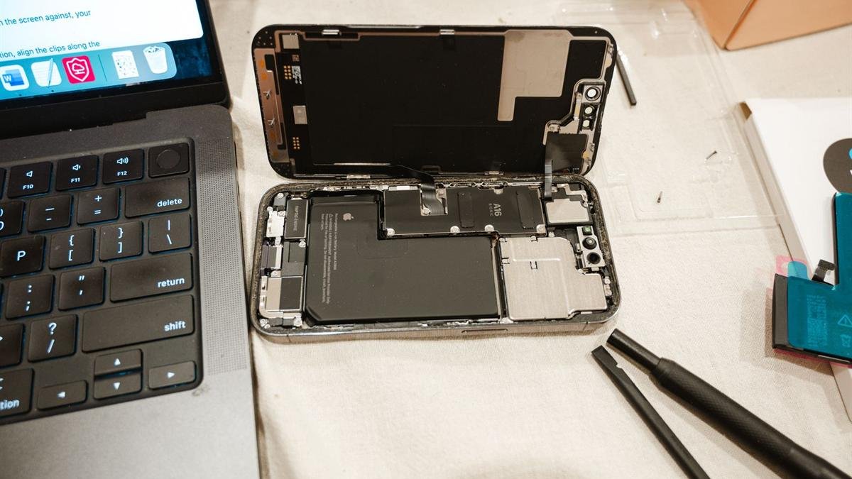 An iPhone opened up on a table with its battery visible, next to repair tools and a laptop