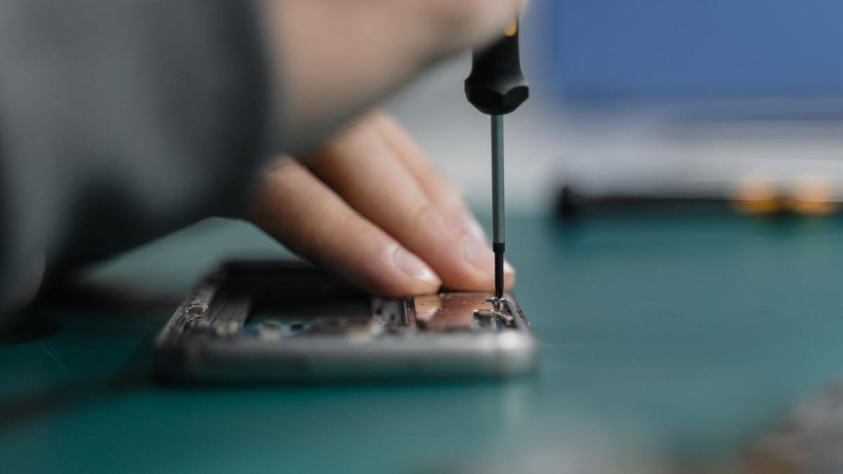 A hand holding a small screwdriver over the open back of a phone on a green repair mat