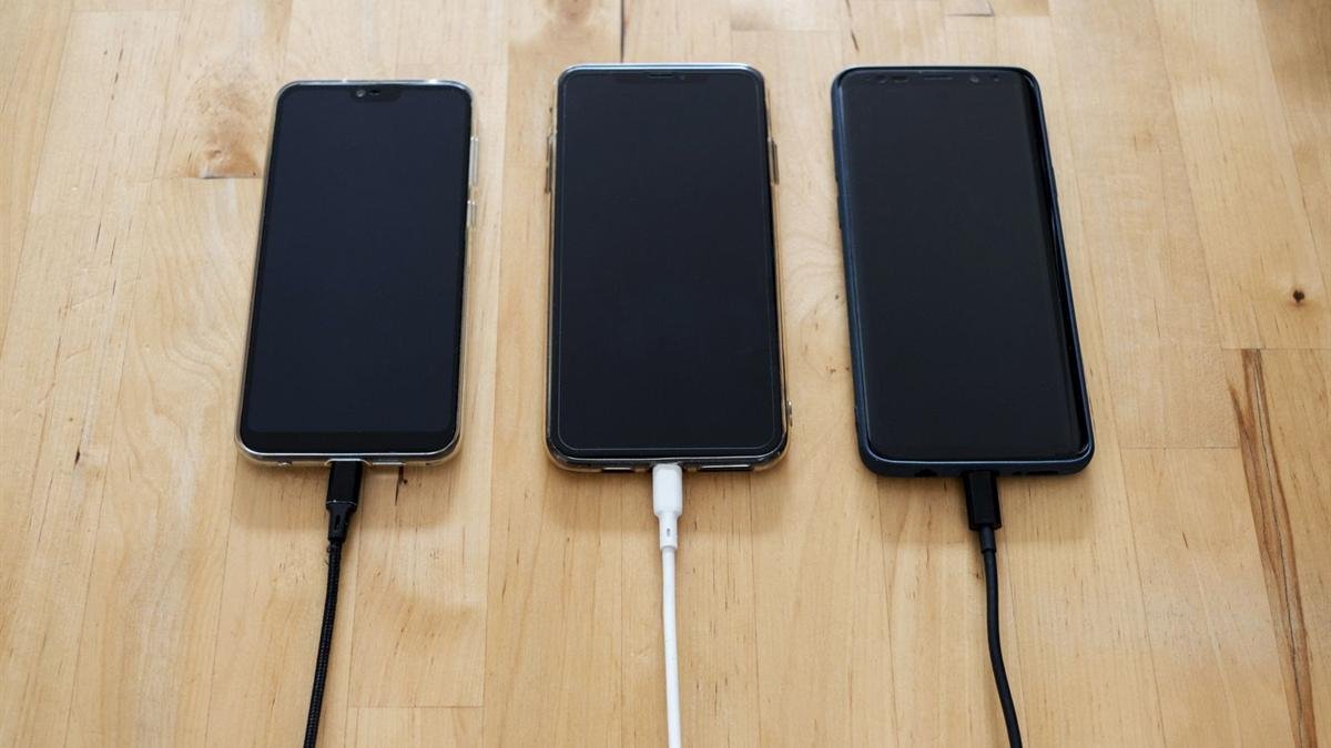 Three smartphones lying on a wooden table, each connected to a charging cable