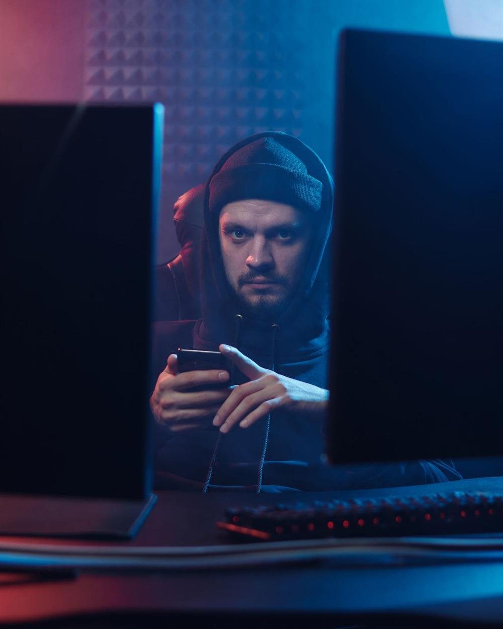 A person in a dark hooded top holding a smartphone in front of two computer monitors