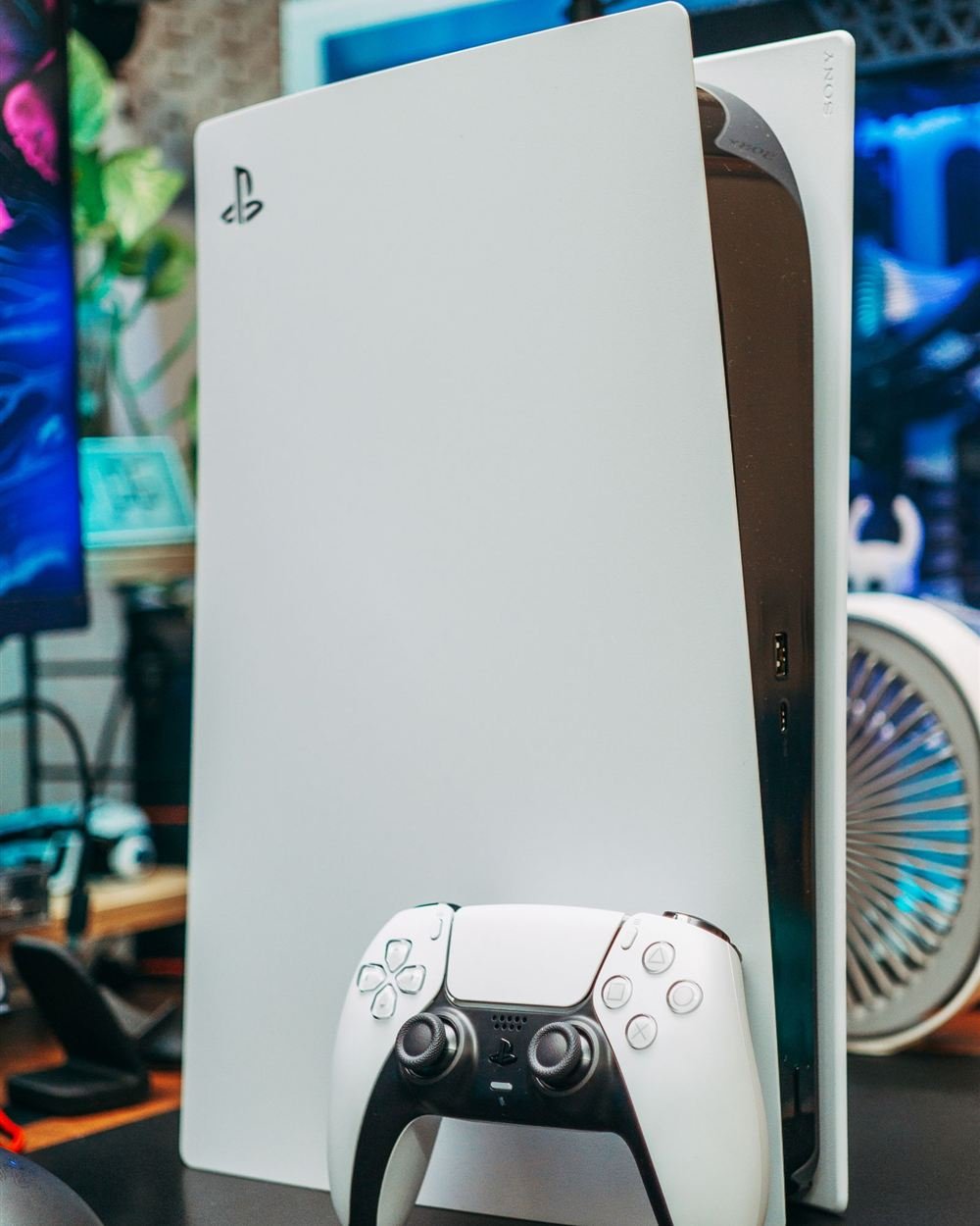 A white PS5 console standing upright on a desk with a controller in front of it and a small desk fan in the background