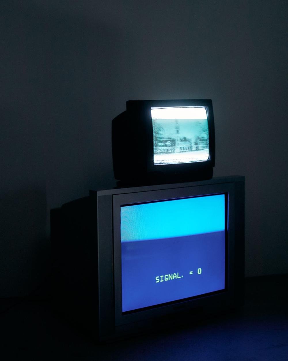 A small old TV on top of another showing a blue screen with the words SIGNAL equals 0 in a dark room
