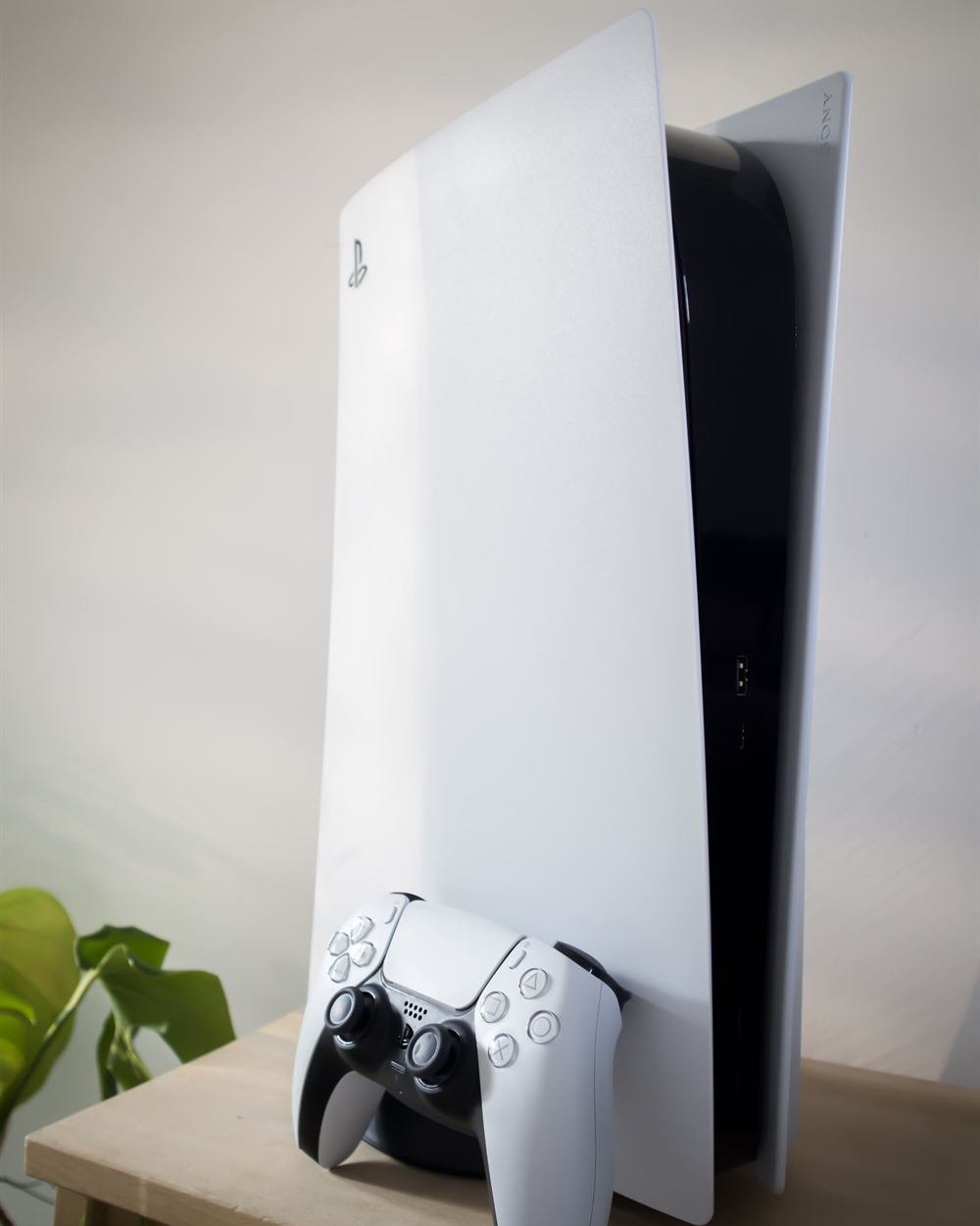 A white PS5 console standing upright on a wooden shelf with a DualSense controller in front of it