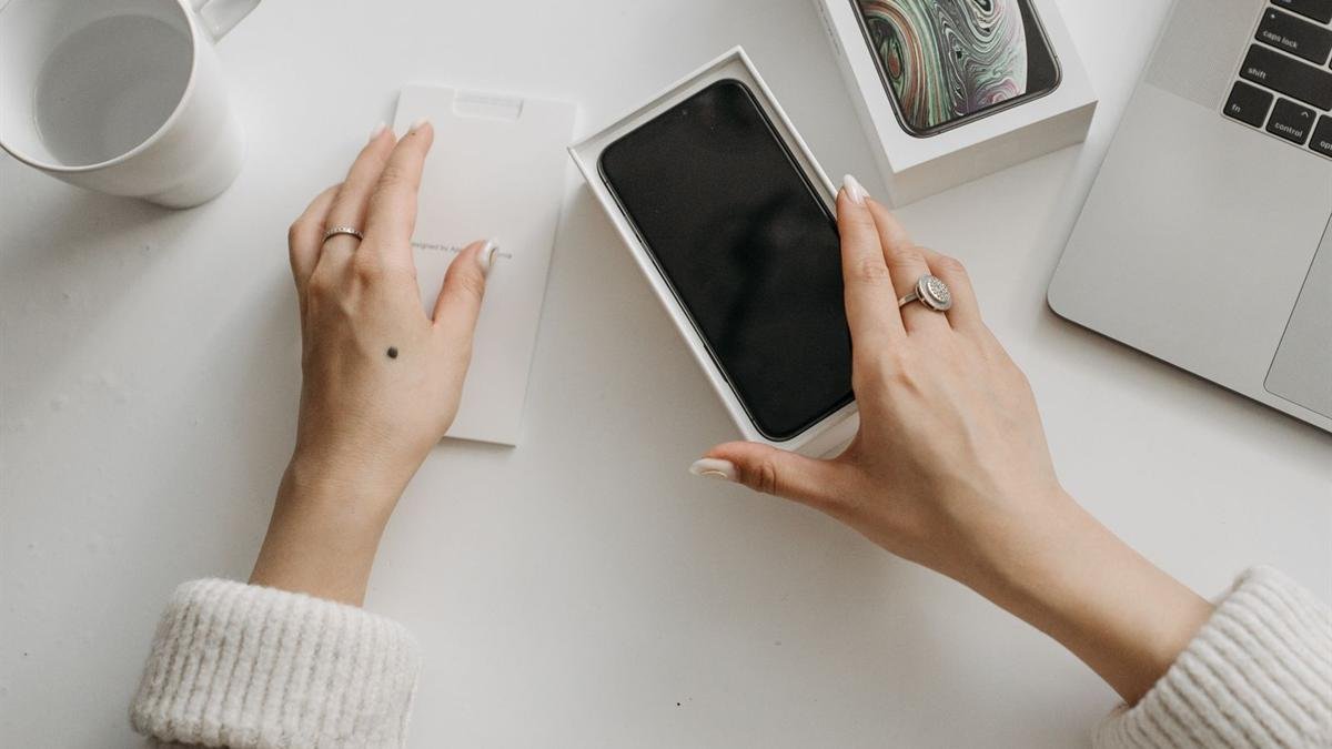 Hands holding a phone in its box on a white desk next to a laptop