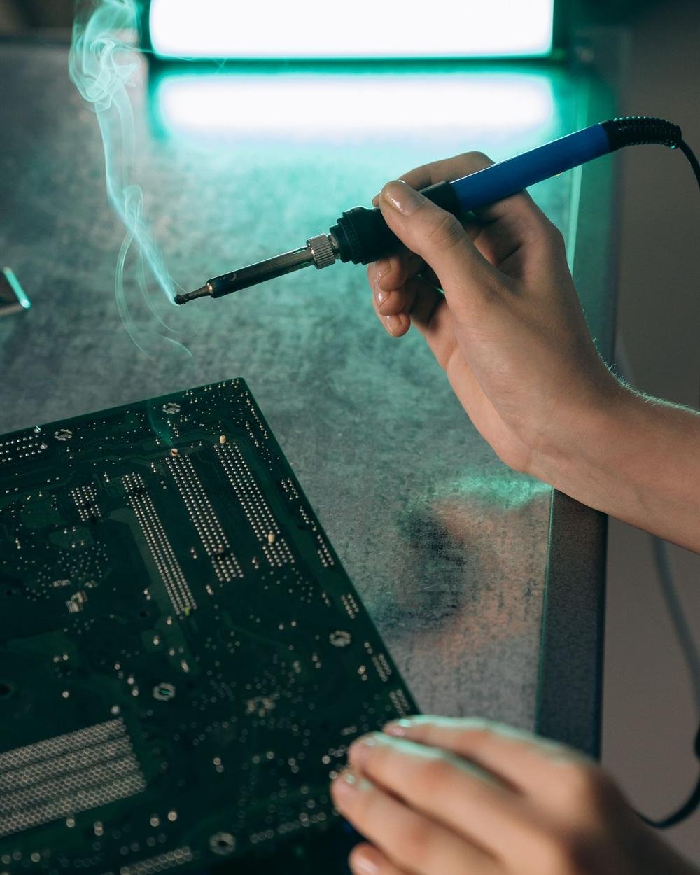 A hand holding a soldering iron with smoke rising over a circuit board lit in green