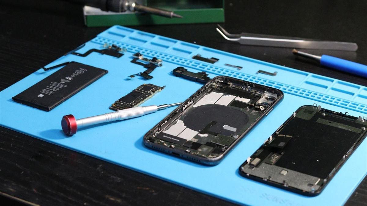 A smartphone taken apart on a blue repair mat, with the battery, logic board and screwdriver laid out beside it