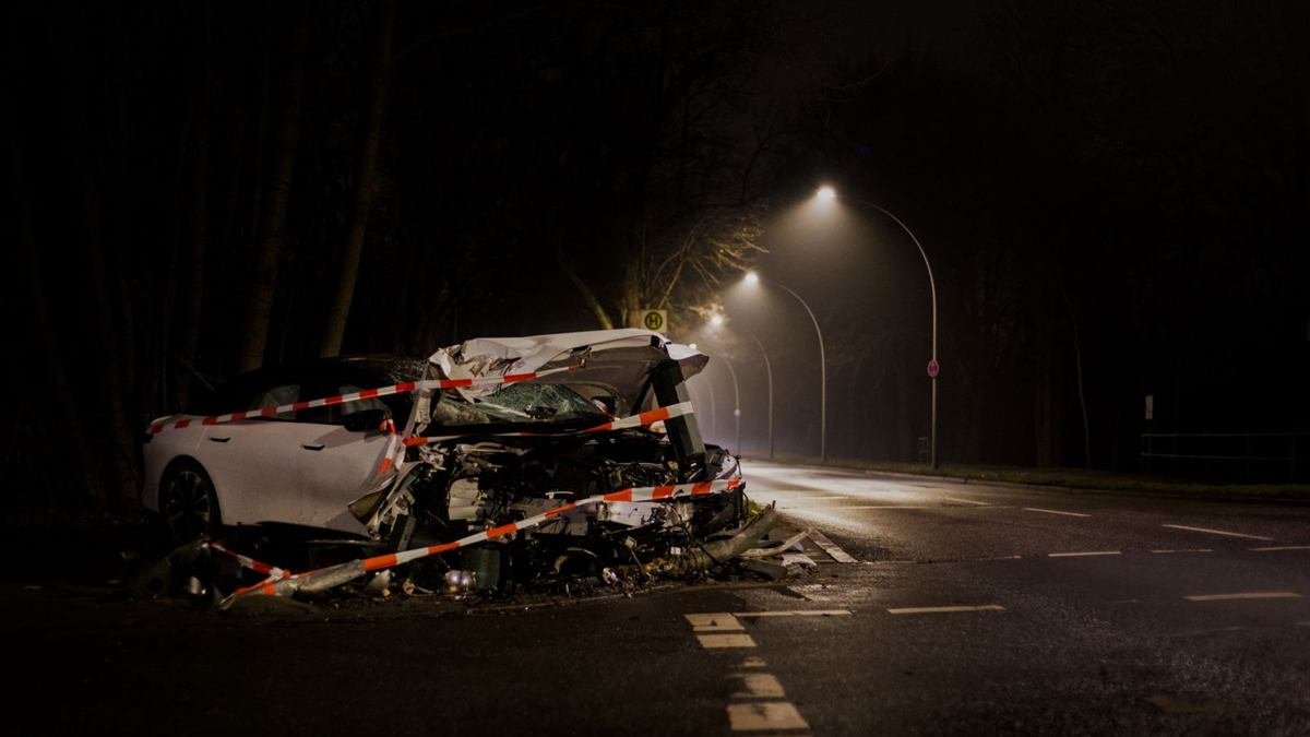 A modern white car with its front end completely destroyed, surrounded by red and white police tape on a dark road at night