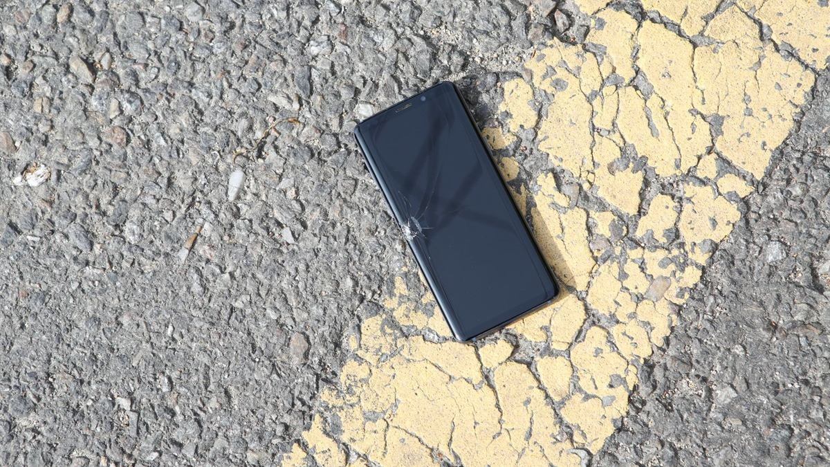 A dark smartphone with a small crack in the corner of its screen, lying on cracked asphalt